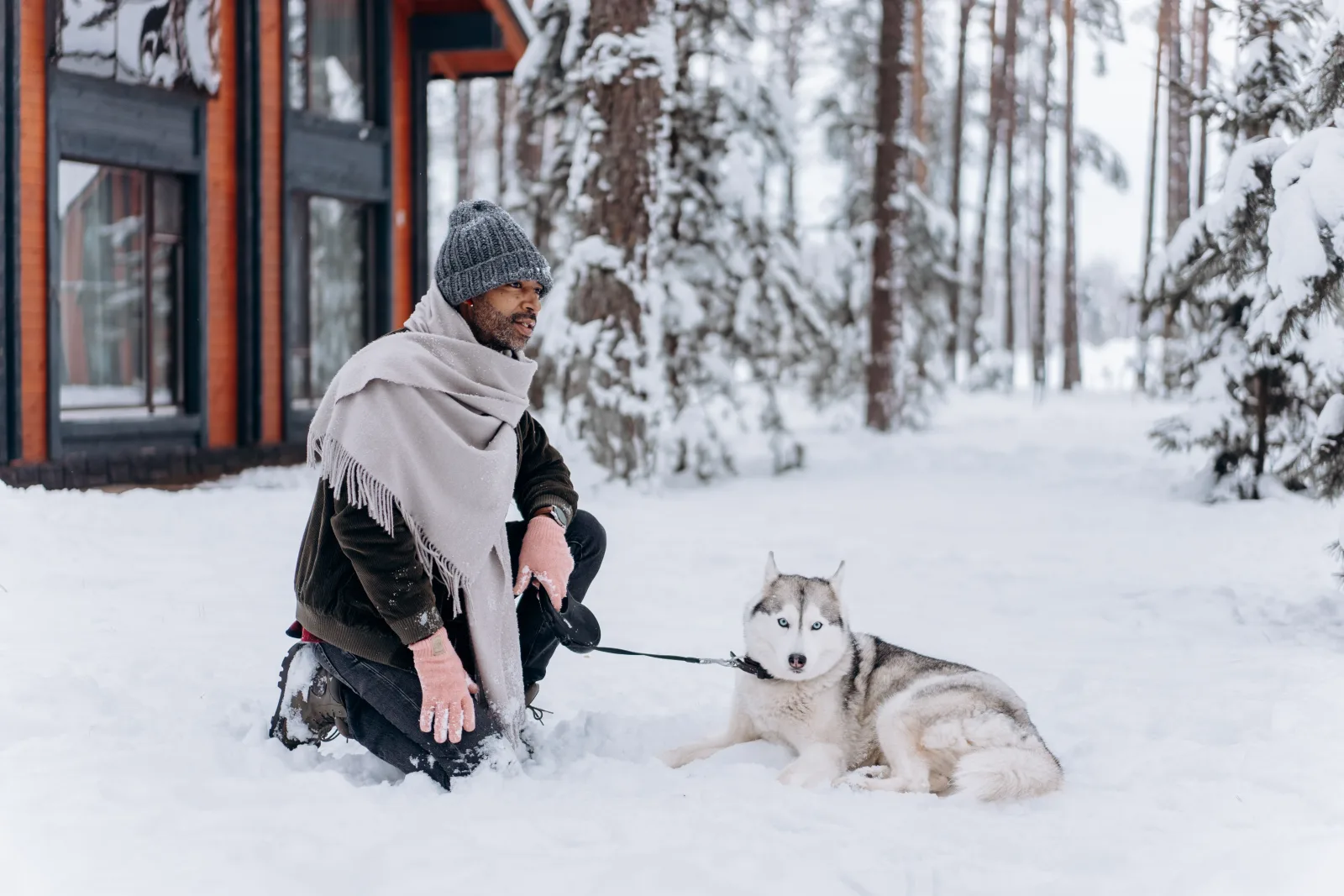 Man kneeling with his leashed husky outside a wooden cabin in a snowy forest